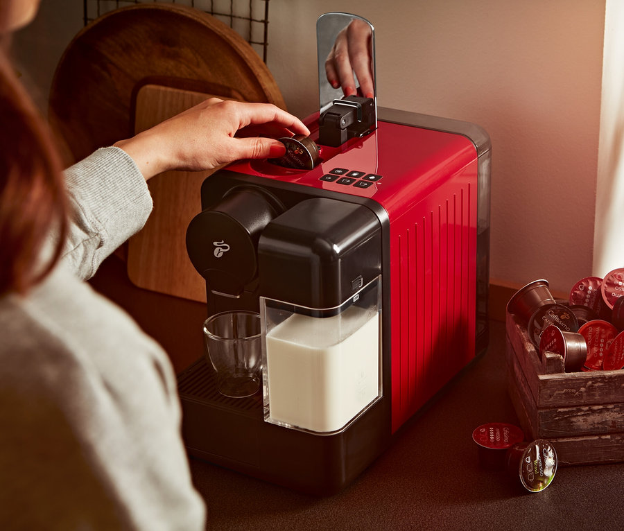 Frau legt Kapsel in rote Cafissimo milk Kaffeemaschine. Daneben stehen ein Glas Milch und eine Holzkiste mit Kapseln.