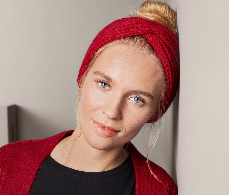 Blonde Frau lehnt an der Wand und trägt ein rotes Strickstirnband.