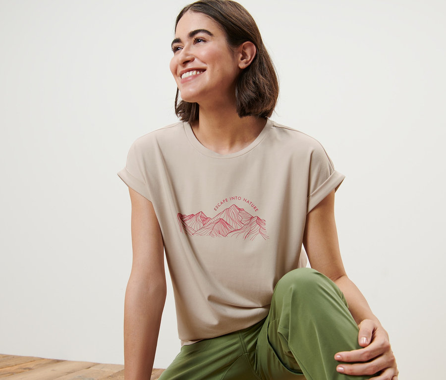 Eine Frau sitzt auf einem Holzboden und trägt ein beiges T-Shirt mit einem Bergaufdruck.