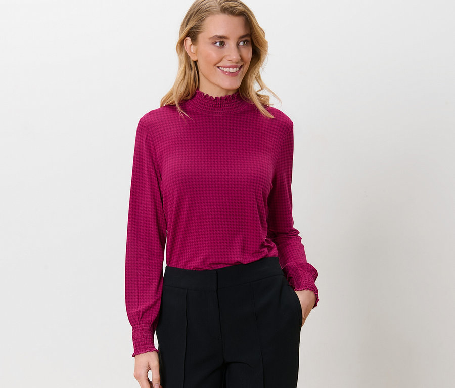 Frau trägt ein bedrucktes Blusenshirt mit Stehkragen und eine schwarze Hose.