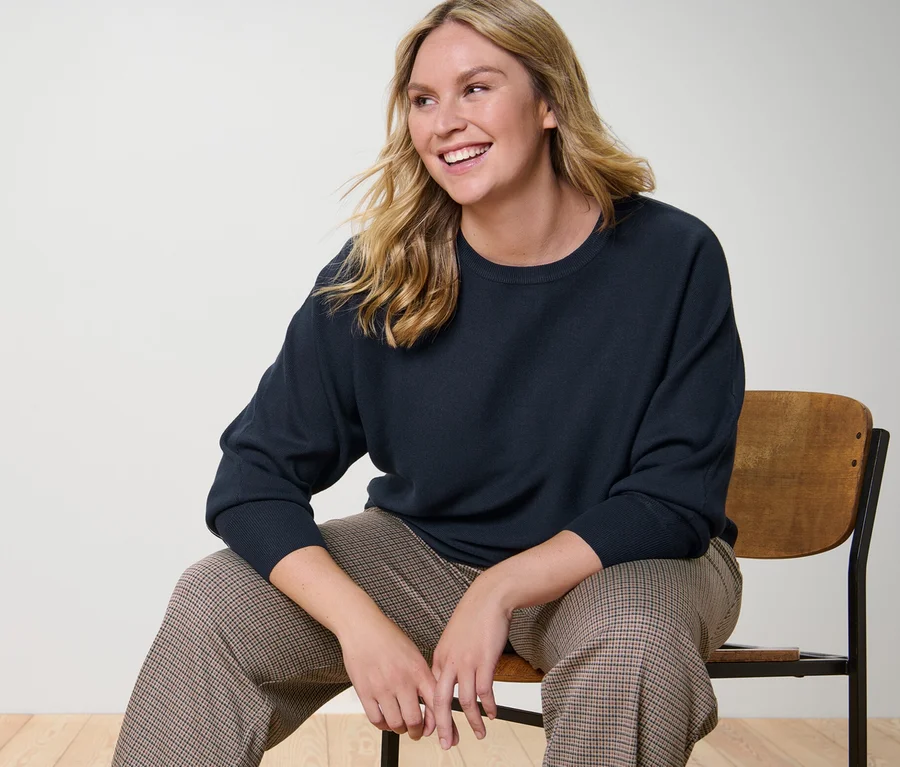 Frau sitzt auf einem Stuhl und trägt einen marineblauen Feinstrickpullover mit Fledermausarm und eine bequeme Jacquardhose.