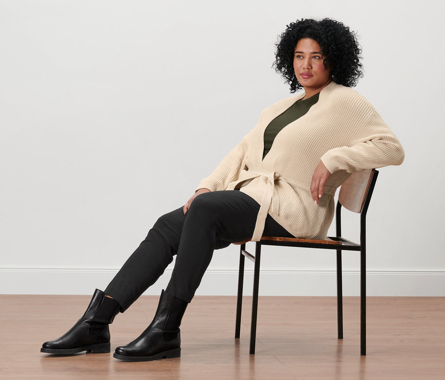 Eine Frau sitzt auf einem Stuhl und trägt einen Wickel-Cardigan, ein olivgrünes Shirt mit 3/4-Arm, eine schwarze Stretchhose und Lederboots.