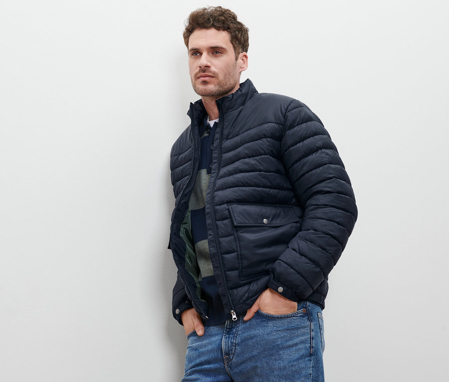 Mann lehnt an Wand, trägt marineblaue Steppjacke und Jeans.