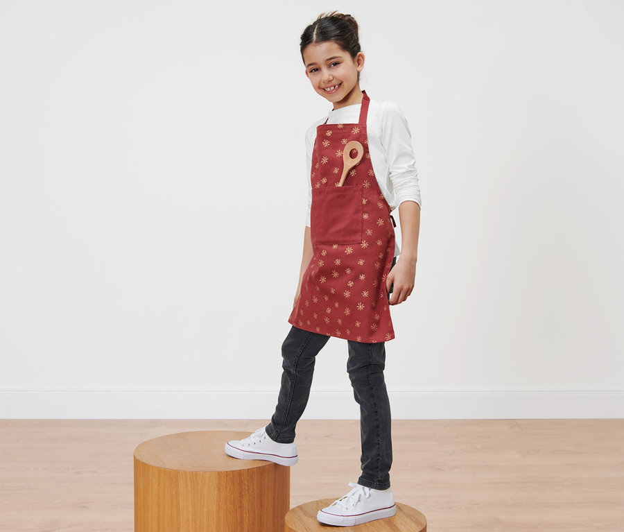 Mädchen steht auf Holzpodest und trägt eine rote Kinderschürze mit Schneeflocken.