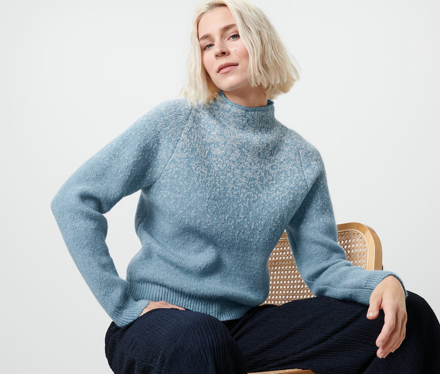 Blonde Frau sitzt auf einem Stuhl und trägt einen blauen Pullover mit Glitzerstrick.