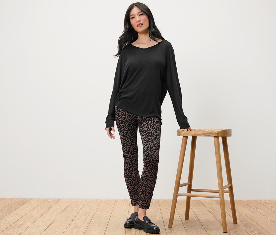 Frau steht neben einem Holzhocker und trägt ein schwarzes Langarmshirt und bedruckte Heavy-Leggings.