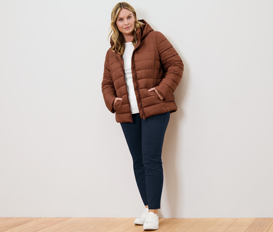 Frau posiert in cognacfarbener Steppjacke mit Kapuze, dunkelblauer Stretchhose und weißem Shirt mit 3/4-Arm.