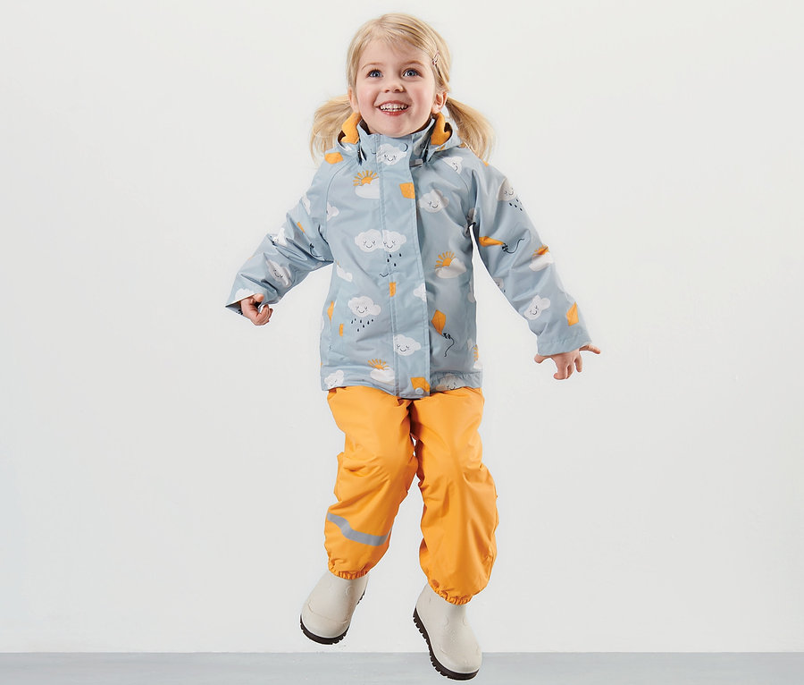 Mädchen springt vor grauem Hintergrund und trägt eine hellblaue Kleinkind-Thermo-Regenjacke mit Alloverprint und orangefarbene Regenhose.