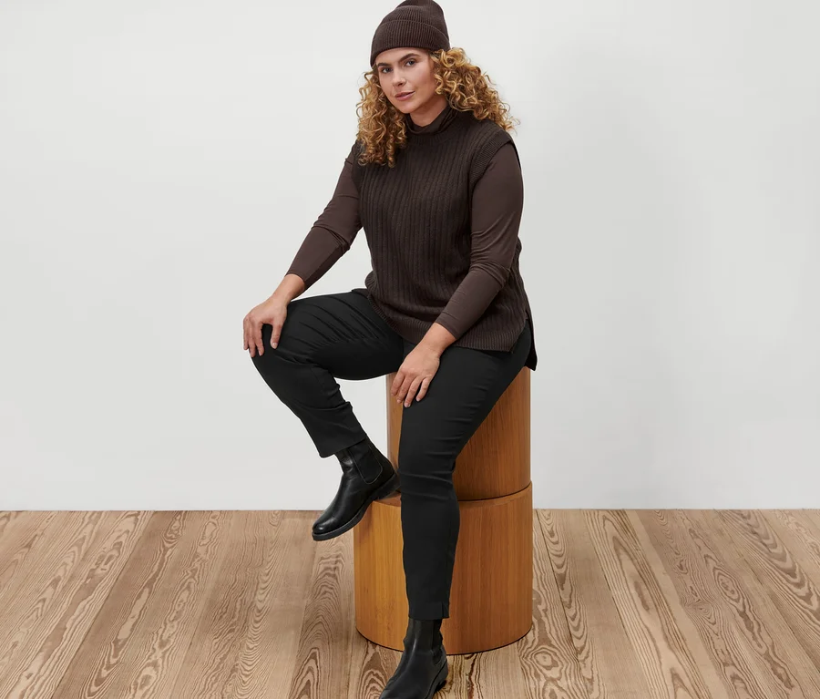 Eine Frau sitzt auf einem Holzhocker und trägt ein braunes Rollkragenshirt, einen Strick-Pullunder, eine braune Strickmütze und eine schwarze Stretchhose.