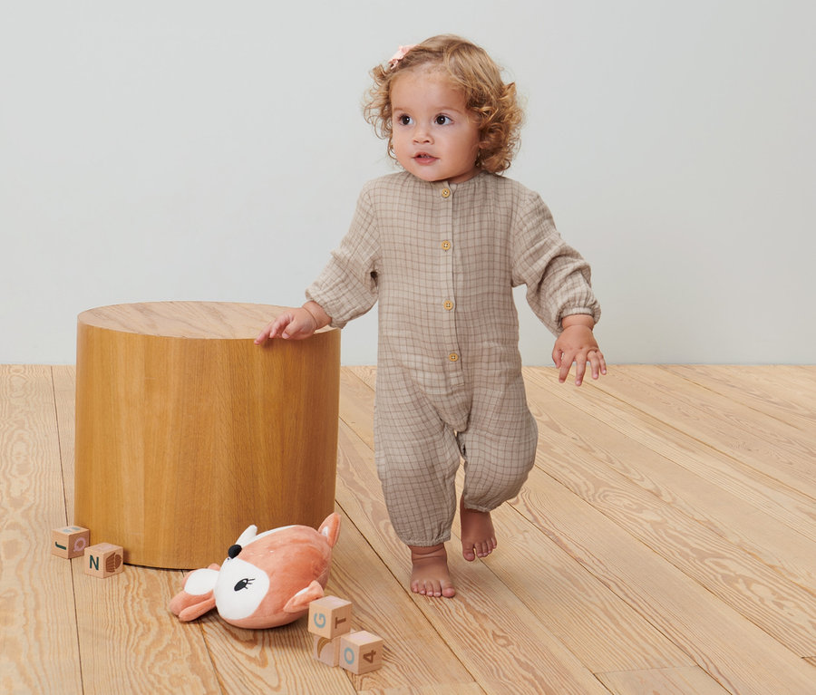 Kleinkind in Baby-Strampler steht auf Holzboden neben Holzhocker und Plüschtier.