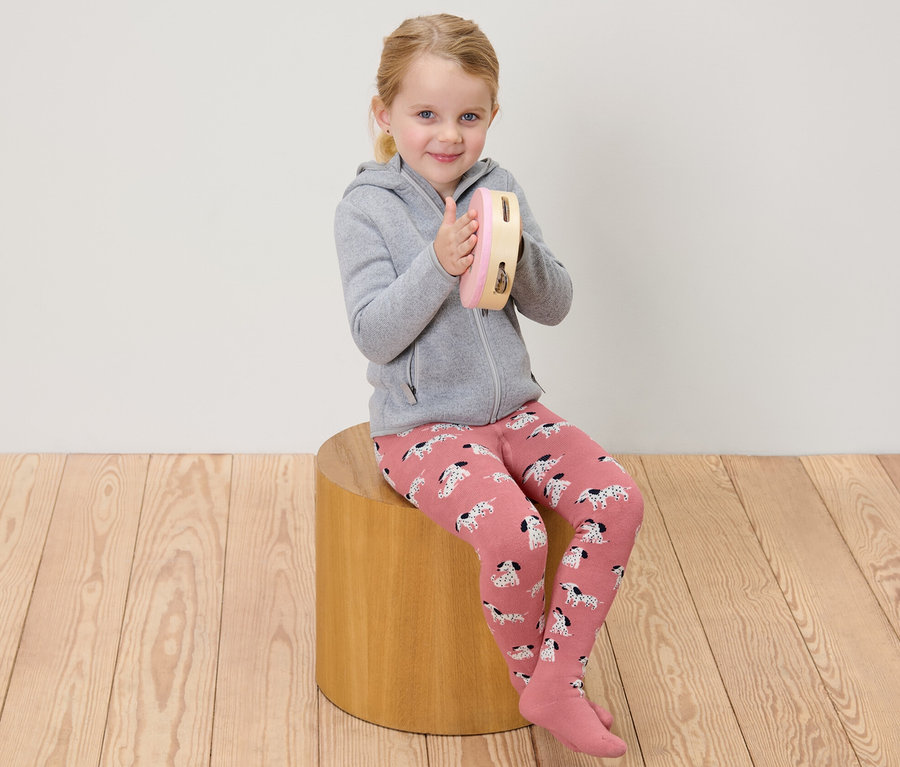 Mädchen sitzt und spielt auf einem Tamburin, trägt rosa und graue Kinder-Thermostrumpfhosen.