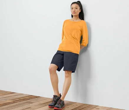 Eine Frau lehnt an einer Wand und trägt ein oranges Funktionsshirt mit 3/4-Ärmeln, schwarze Shorts und Schuhe.