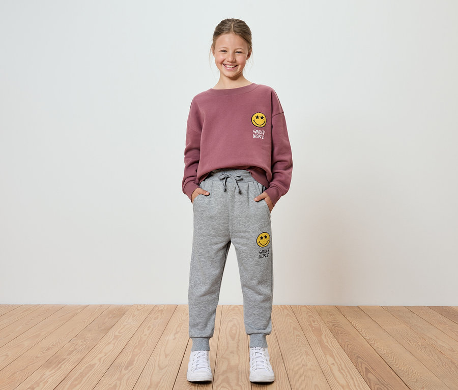 Mädchen in bordeauxfarbenem Sweatshirt und grauer Jogginghose mit Smiley steht vor einer weißen Wand.