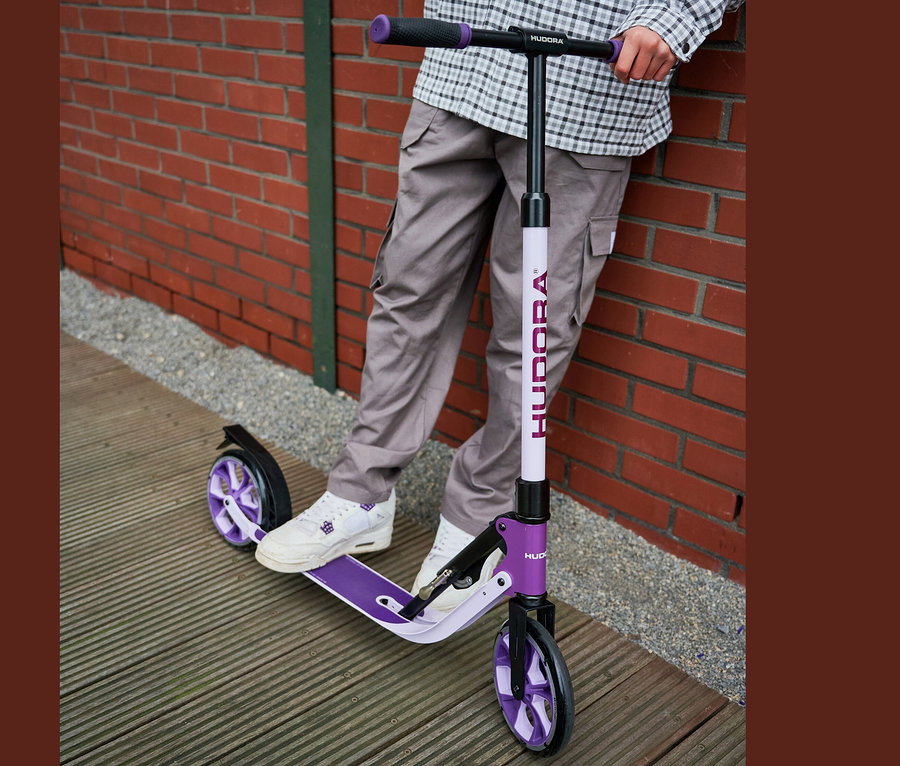 Mann steht mit lavendelfarbenem HUDORA Scooter BigWheel® 205 an Ziegelwand.