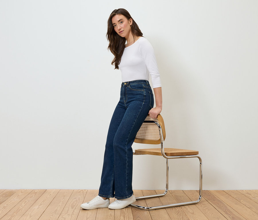 Frau sitzt seitlich auf einem Stuhl, trägt blaue Jeans und ein weißes T-Shirt.