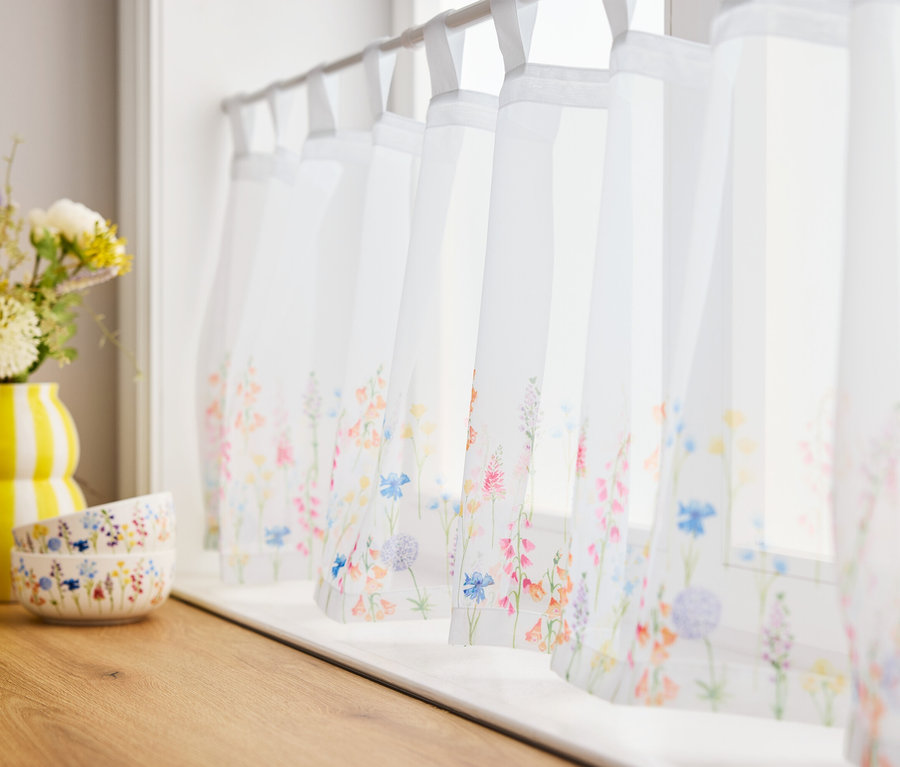 Blumen in gelber Vase und zwei Schalen auf Holzbrett vor Fenstern mit Blumenvorhängen.