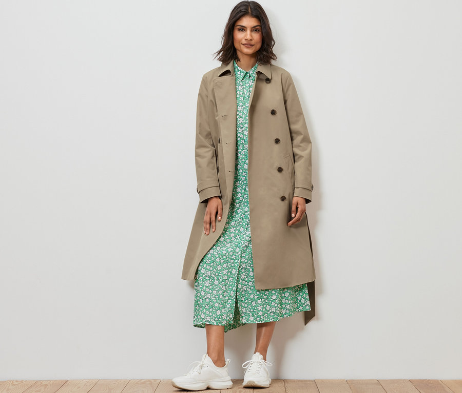 Frau steht und trägt einen Trenchcoat in Sand.