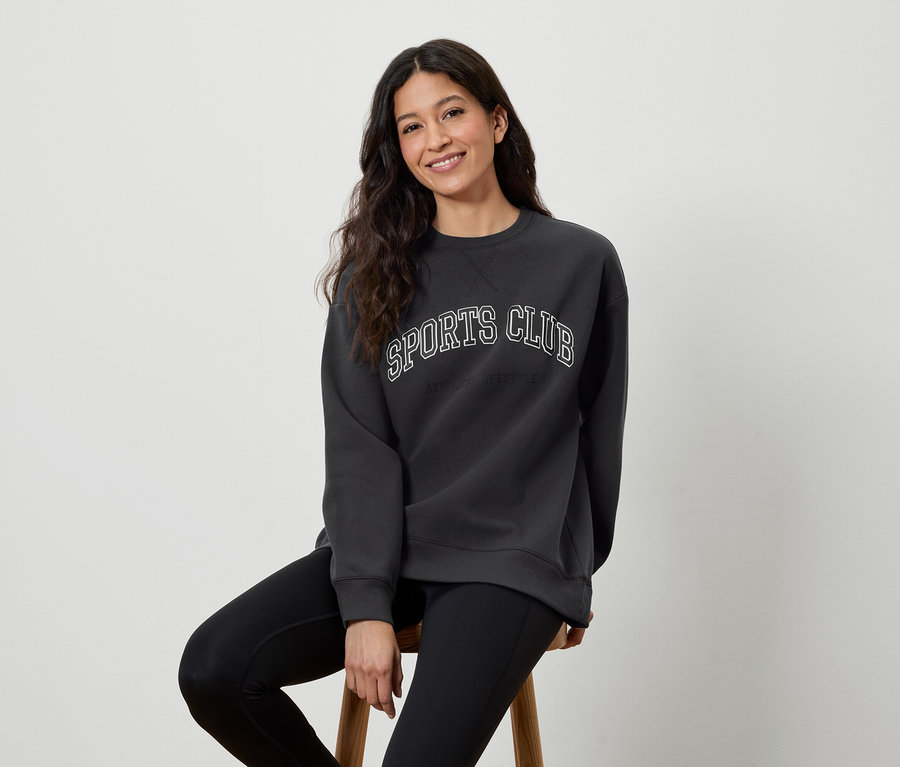 Frau sitzt auf einem Hocker, trägt ein dunkelgraues Sweatshirt mit der Aufschrift SPORTS CLUB und schwarze Leggings.
