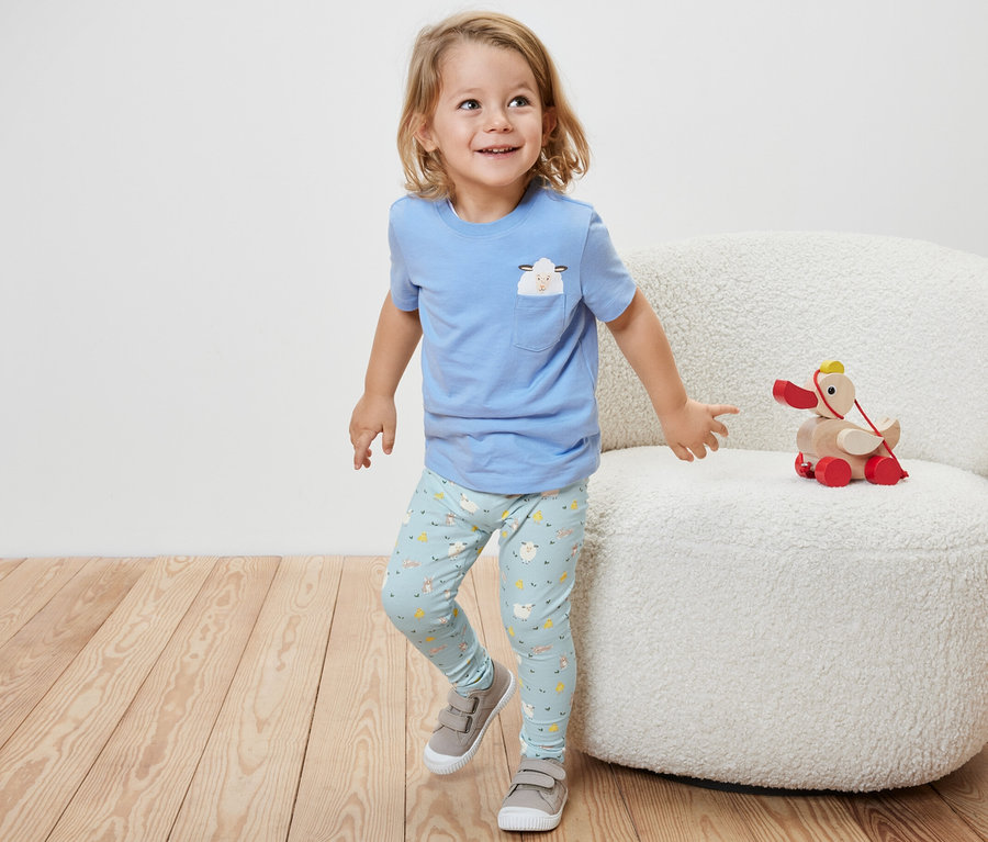Ein kleiner Junge in einem blauen Kurzarmshirt und 2 Kinder-Leggings, blau, steht neben einem weißen Sessel, auf dem ein Holzspielzeug sitzt.