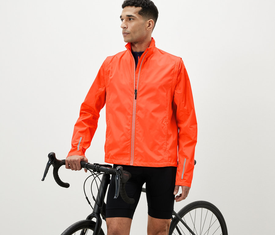 Mann steht mit einem Fahrrad und trägt eine orangefarbene 2-in-1-Fahrradjacke.
