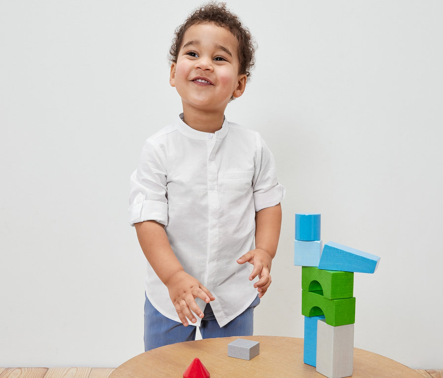Ein Junge in einem Kinder-Hemd spielt mit bunten Holzbausteinen auf einem Tisch.