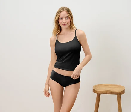 Blonde Frau posiert in schwarzem Top und Unterhose vor einem Holzhocker.
