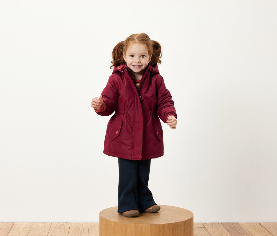 Ein Mädchen mit roten Zöpfen steht auf einem Holzsockel und trägt eine burgunderrote Jacke und dunkelblaue Hosen.