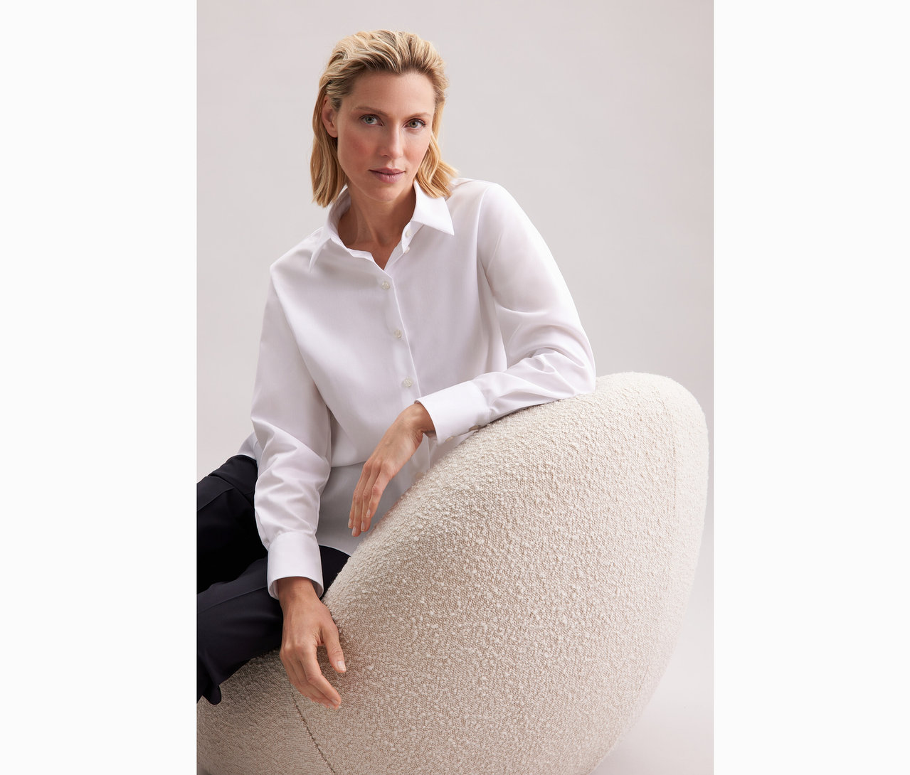 Frau sitzt auf einem Sessel und trägt eine weiße Seidensticker Oversize Longbluse.