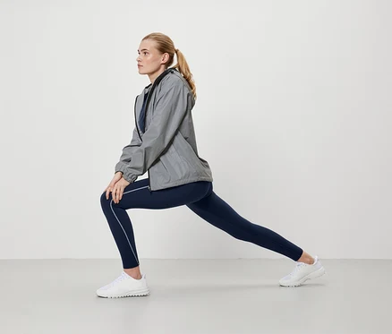 Blonde Frau in grauer Jacke und blauen Leggings dehnt sich.