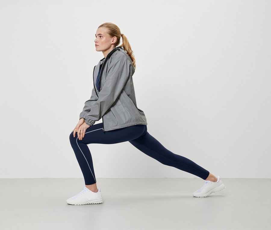 Blonde Frau in grauer Jacke und blauen Leggings dehnt sich.