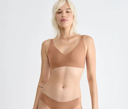 Blonde Frau trägt einen sloggi ZERO Feel 2.0 Soft Bra in Nostalgic Brown und passende Unterwäsche, blickt nach vorne. Sie hat ein Tattoo am linken Unterarm.