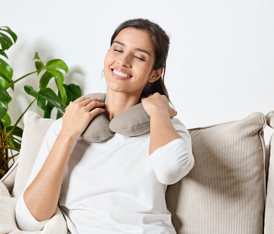 Frau sitzt mit geschlossenen Augen auf einem Sofa und hat ein graues Nackenkissen um den Hals.