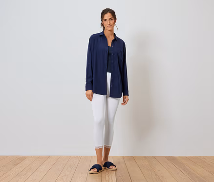 Frau steht vor einer weißen Wand und trägt ein blaues Hemd, weiße Leggings und blaue Hausschuhe.
