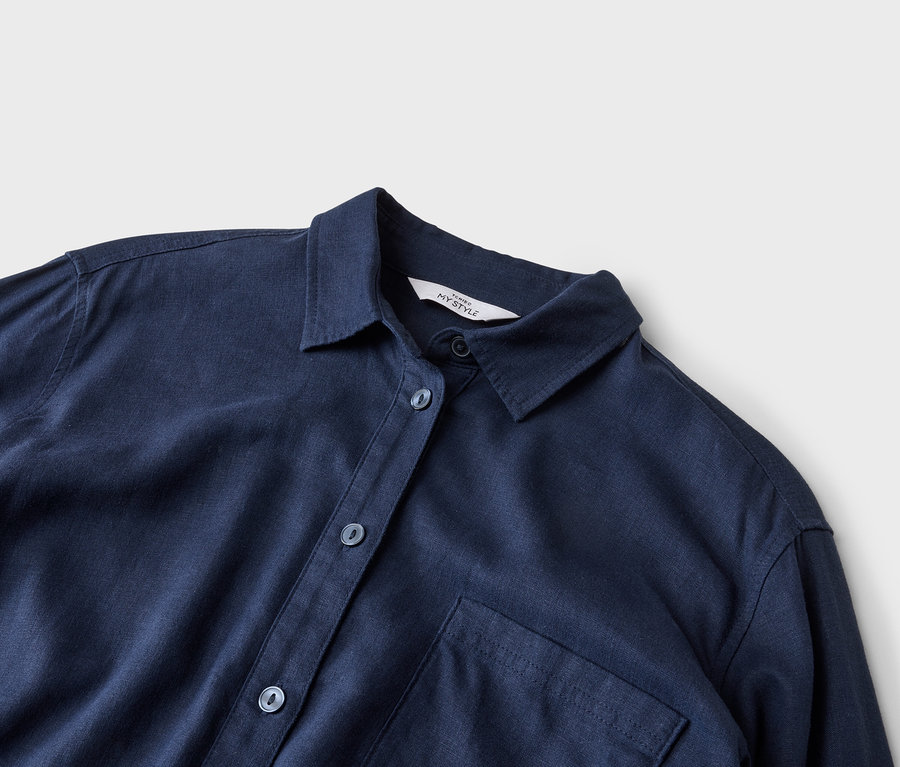 Nahaufnahme eines marineblauen Hemdes mit Kragen, Knöpfen und einer einzelnen Brusttasche auf neutralem Hintergrund.