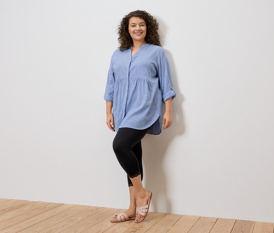 Frau steht in blauem Hemd, schwarzen Leggings mit Spitze und rosa Sandalen vor einer weißen Wand.