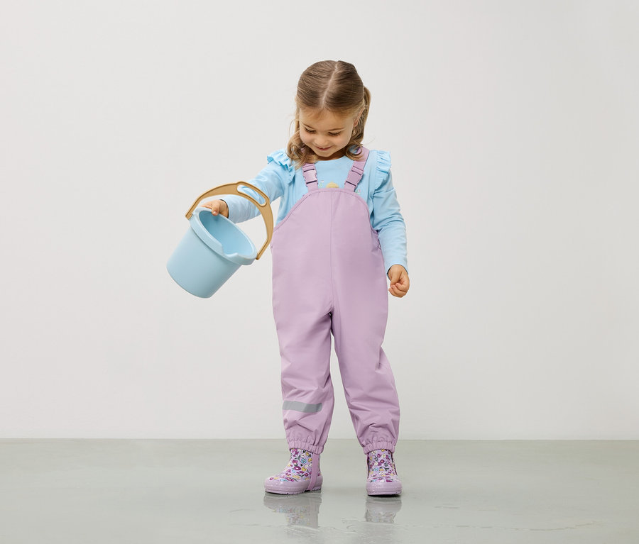 Mädchen mit hellblauer Bluse und lila Overall hält einen hellblauen Eimer.