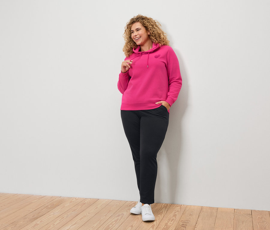 Frau mit lockigen Haaren, die einen pinkfarbenen Kapuzenpullover und schwarze Hosen trägt.