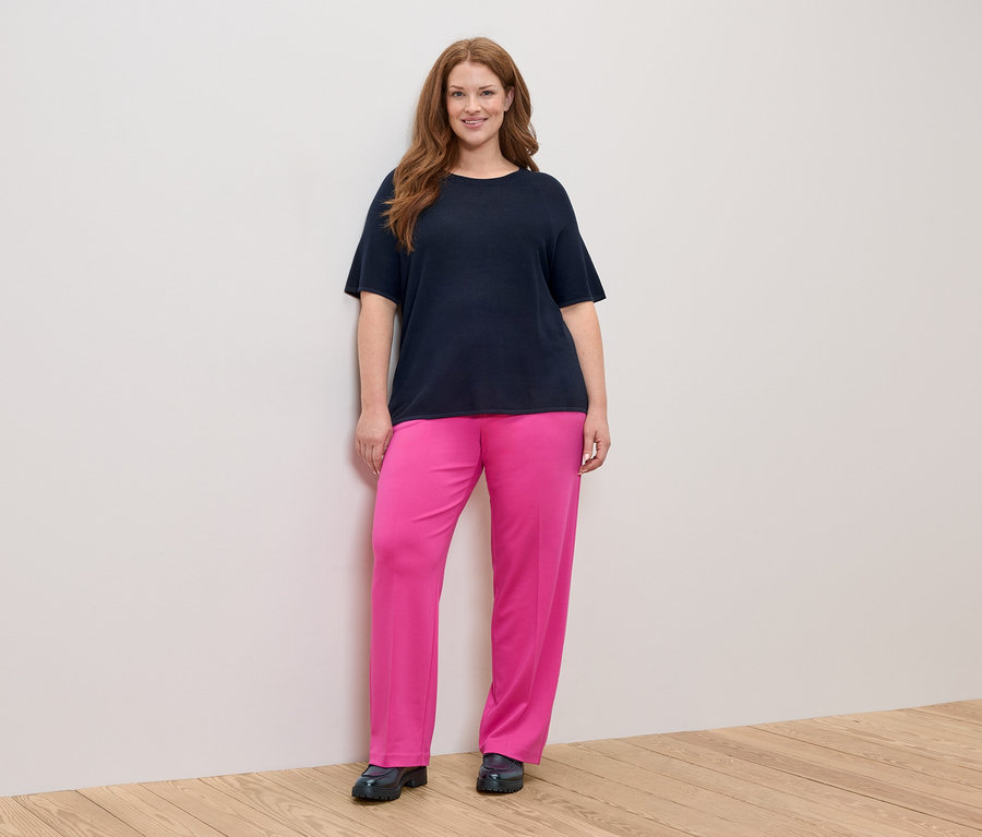 Frau mit langen, roten Haaren, die ein marineblaues T-Shirt und eine pinke Hose trägt.