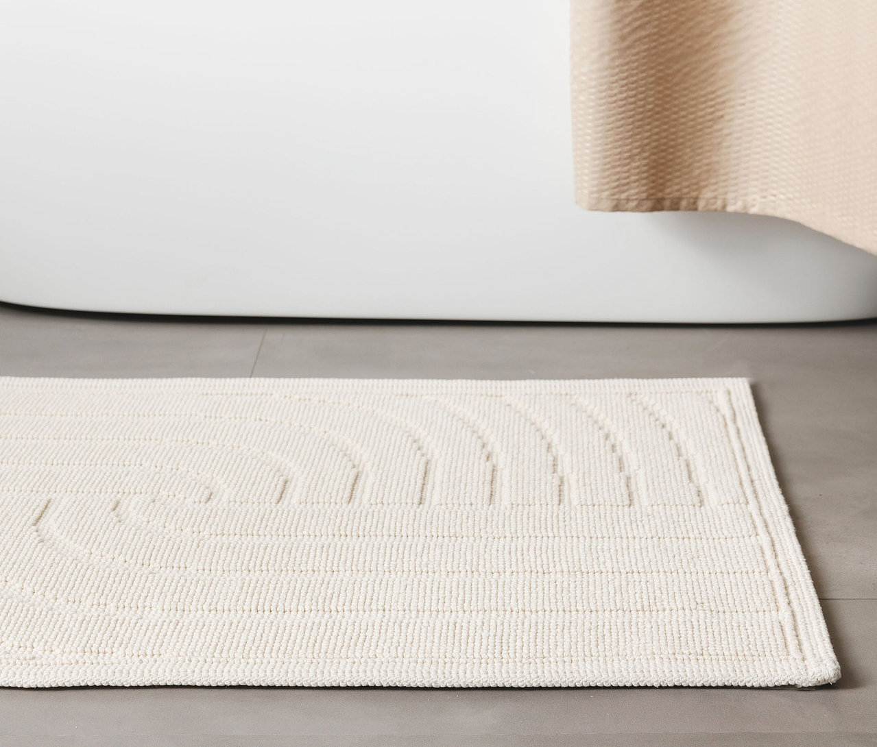 Eine cremefarbene, rechteckige Badematte »Jojo« von benuta mit Wellenmuster liegt vor einer weißen Badewanne.