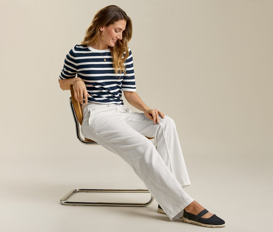 Frau sitzt auf Stuhl und trägt Wide-Komfort-Jeans, gestreiftes Shirt und flache Schuhe.