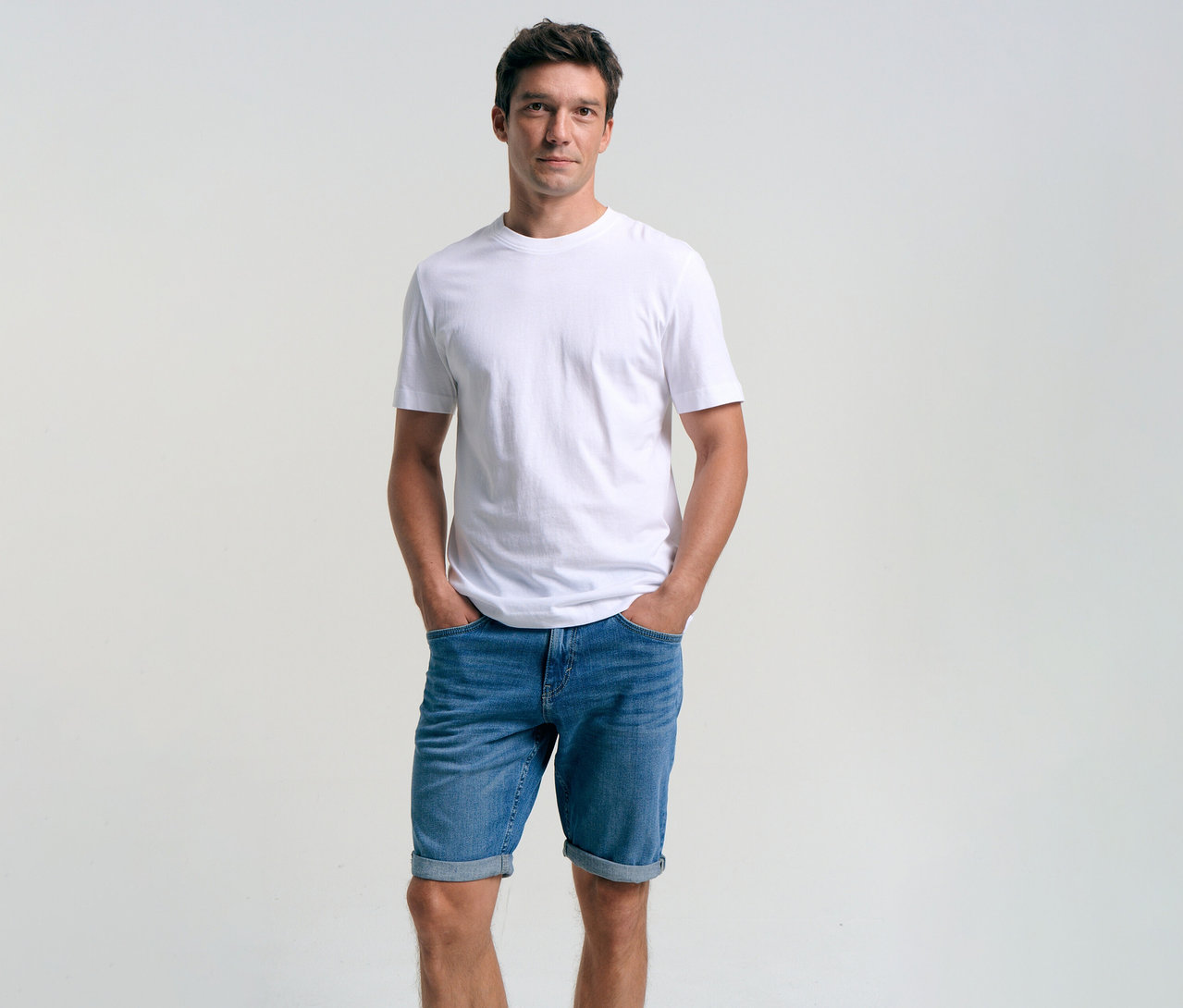 Mann mit weißem T-Shirt und Tom Tailor TTJOSH REGULAR SLIM Jeansshorts in Mid Stone Bright Blue Denim mit Händen in den Hosentaschen.
