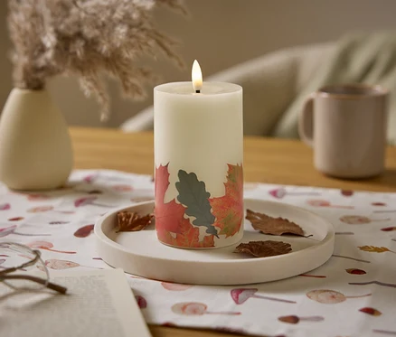 Eine brennende, weiße Kerze mit Blättermuster steht auf einem Tisch mit einem beige farbenen Krug und einer Vase.