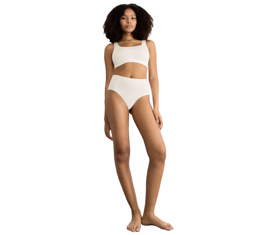 Frau mit Locken trägt ein seidenweißes Bralette mit eckigem Ausschnitt und passende sloggi ZERO Feel 2.0 High Waist Höschen, barfuß stehend.
