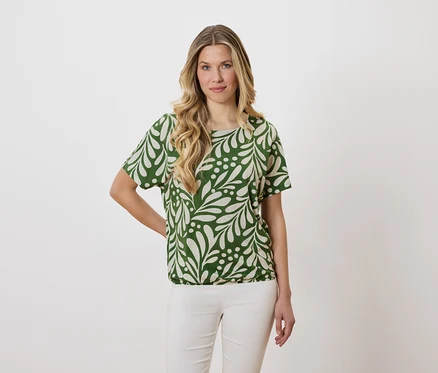 Frau mit blonden, gewellten Haaren trägt eine grüne Bluse mit Blattmuster und weiße Hosen.
