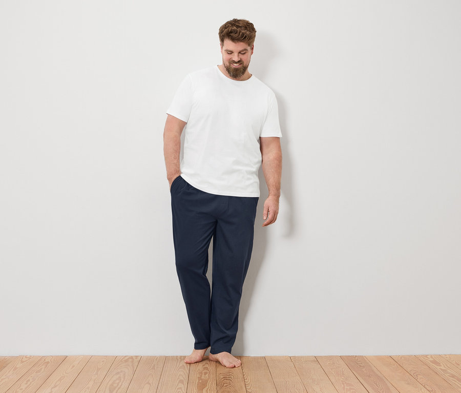 Mann steht barfuß mit der Hand in der Hosentasche vor einer weißen Wand.