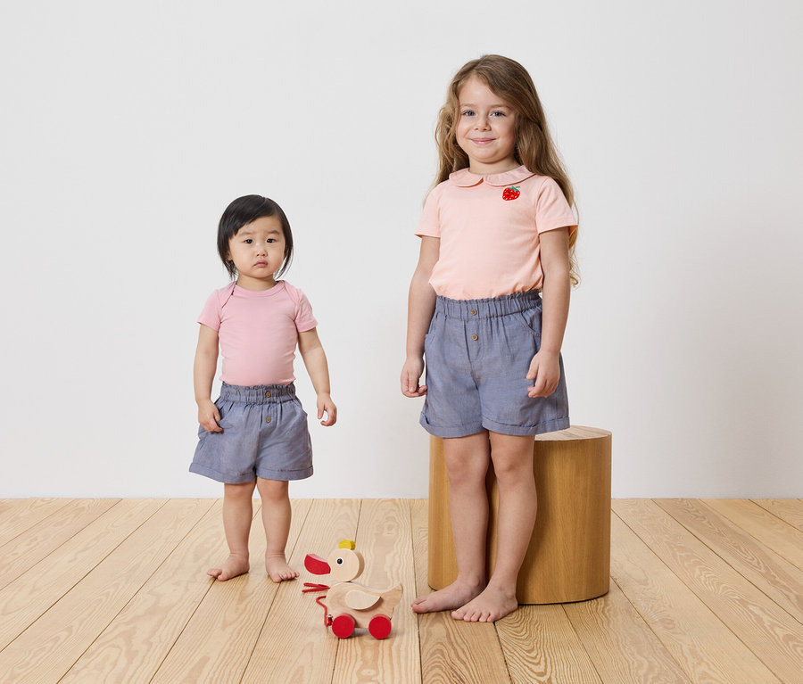 Zwei Mädchen stehen auf einem Holzboden, beide tragen rosa Oberteile und blaue Shorts. Ein Holzspielzeug befindet sich auf dem Boden.
