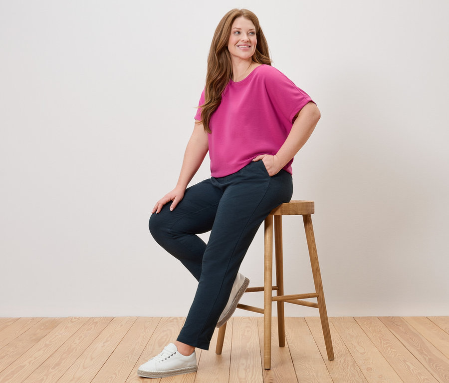 Frau sitzt auf einem Hocker, trägt ein pinkes Oberteil, eine blaue Hose und weiße Schuhe.