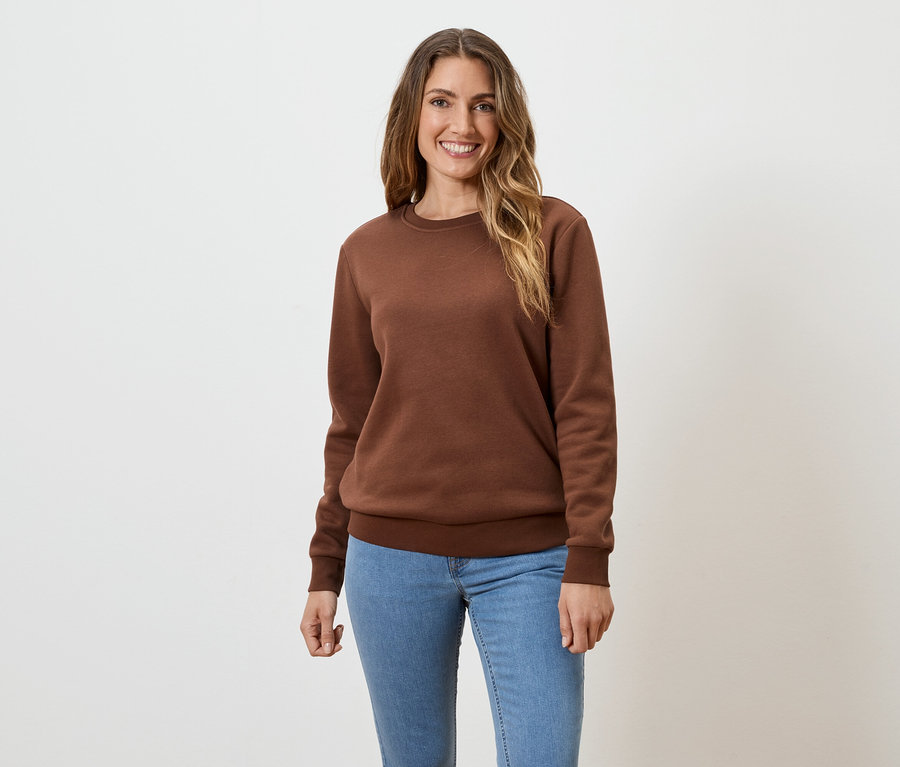 Frau mit langen, gewellten, braunen Haaren, die ein braunes Sweatshirt und Jeans vor einem weißen Hintergrund trägt.