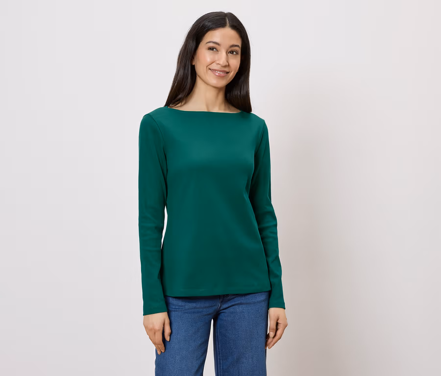 Frau trägt ein grünes Langarmshirt und Jeans vor einem weißen Hintergrund.