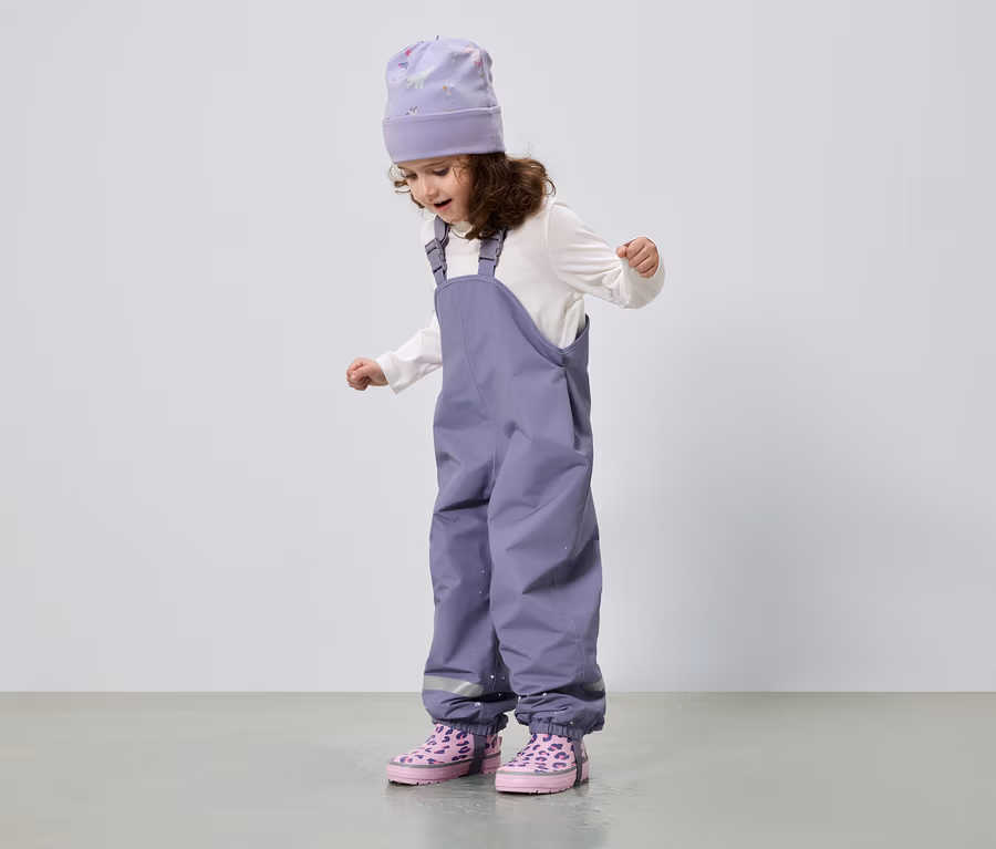 Ein kleines Mädchen mit lockigem Haar trägt eine lila Mütze, eine weiße Bluse, eine lila Hose und rosa Stiefel.